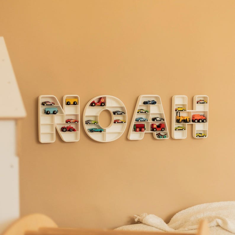 K&ouml;nnte beinhalten: Eine an der Wand befestigte, cremefarbene Holznamensanzeige mit der Aufschrift "NOAH". Jeder Buchstabe ist ein Regal, das mit Miniatur-Spielzeugautos in verschiedenen Farben gef&uuml;llt ist. Die Anzeige ist an einer hellbraunen Wand angebracht.