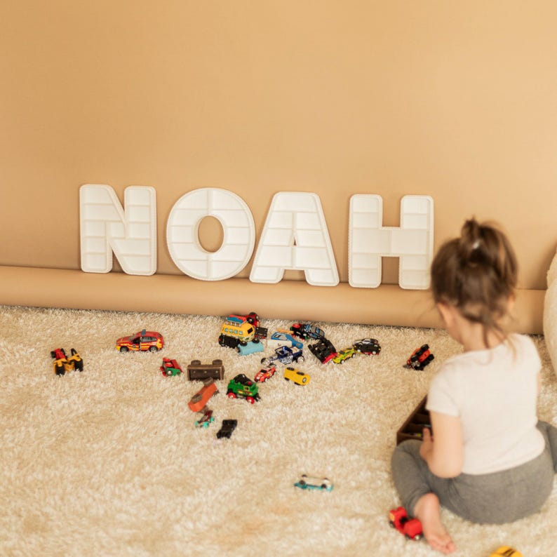 K&ouml;nnte beinhalten: Wei&szlig;e Buchstaben im Blockstil mit der Aufschrift "NOAH" sind an einer beigefarbenen Wand &uuml;ber einem flauschigen, cremefarbenen Teppich angebracht. Eine Sammlung von Spielzeugautos in verschiedenen Farben ist auf dem Teppich verstreut. Ein junges Kind spielt mit den Spielzeugautos.