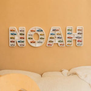 K&ouml;nnte beinhalten: Wei&szlig;e, an der Wand befestigte Regale bilden den Namen "NOAH", jeder Buchstabe gef&uuml;llt mit bunten Spielzeugautos. Die Regale sind an einer hellorangen Wand angebracht und schaffen eine spielerische Pr&auml;sentation f&uuml;r Spielzeugautos.