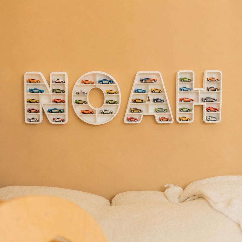 K&ouml;nnte beinhalten: Wei&szlig;e, an der Wand befestigte Regale bilden den Namen "NOAH", jeder Buchstabe gef&uuml;llt mit bunten Spielzeugautos. Die Regale sind an einer hellorangen Wand angebracht und schaffen eine spielerische Pr&auml;sentation f&uuml;r Spielzeugautos.