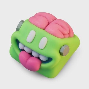 Op de afbeelding: Een grillige, cartoonachtige sculptuur van een groen, vierkant karakter met een grote, open mond en een roze tong die uitsteekt. Het karakter heeft witte tanden, witte ovale ogen en roze hersenachtige uitsteeksels bovenop. Grijze bouten zitten aan de zijkanten.