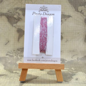 May include: A pink and silver metallic thread spool on a white card with the text "Pirate-Dragon" and "www.facebook.com/piratedragon".