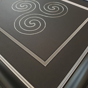 May include: A black framed picture with a white mat and a silver triple spiral design in the center.
