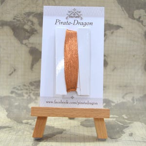 May include: A close-up of a spool of copper-coloured metallic thread on a white card with the text "Pirate-Dragon" and the website address "www.facebook.com/piratedragon".