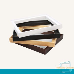 May include: A stack of four rectangular picture frames in varying finishes, including white, black, light wood, and dark wood. The frames are layered on top of each other. The text Frameable Moments is visible in the bottom right corner.