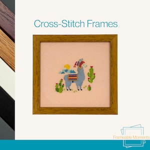 May include: A framed cross-stitch depicting a llama with a colourful blanket, cacti, and mountains. The frame is a warm, light brown colour. The text "Cross-Stitch Frames" is above the frame.
