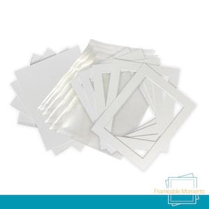 May include: A collection of white rectangular picture frame mounts with central cutouts, accompanied by clear protective plastic sleeves. The set includes multiple stacked paper mounts and transparent bags, all featuring a clean, solid white finish.