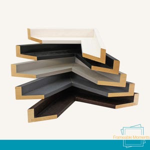 May include: A selection of picture frame corners in shades of white, black, grey, and brown. The frames are made of wood and have painted surfaces. The image is set against a light background.