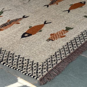 Könnte beinhalten: Ein gewebter Teppich mit beigefarbenem Grund und einem sich wiederholenden Muster aus stilisierten Fischen in Orange-, Braun- und Grüntönen. Der Teppich ist mit einer schwarz-weißen Hahnentrittbordüre und Fransen an der Unterseite versehen.