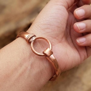 May include: A copper-colored bracelet with a circular clasp is worn on a wrist. The bracelet has a simple, elegant design with a rounded band and a large, circular clasp. The metal has a warm, reddish-brown hue.