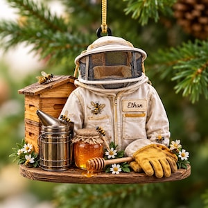 May include: A hanging ornament featuring a beekeeper suit with the name Ethan, a wooden beehive, a metal smoker, a honey jar with a dipper, and yellow gloves. The scene is decorated with small white flowers and several honeybee figures.