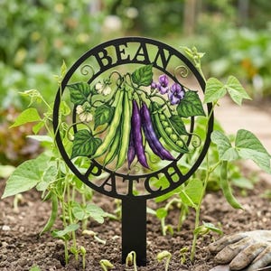 Puede incluir: Un letrero de jardín circular negro con el texto BEAN PATCH en la parte superior e inferior. El centro muestra una ilustración colorida de vainas de frijol verdes y moradas, hojas verdes y pequeñas flores blancas y moradas.