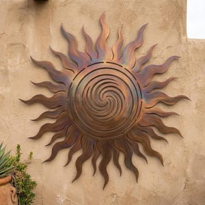 May include: A large, decorative metal sun sculpture with a spiral center and wavy rays. The sun has a bronze finish with hints of blue and purple, mounted on a textured beige wall. A unique piece of outdoor decor.