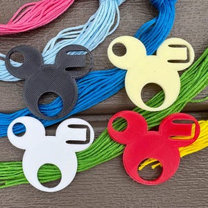 May include: Four plastic Mickey Mouse-shaped cord organizers in black, cream, white, and red, each featuring a central circular cutout and a side slot, arranged over a background of pink, light blue, dark blue, green, and yellow embroidery floss.