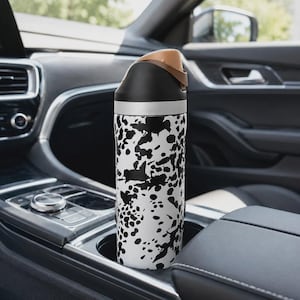 May include: A cylindrical insulated water bottle featuring a white base with an irregular black splatter pattern. The bottle has a black lid with a tan-colored flip-top spout and a silver-toned band separating the lid from the main body.