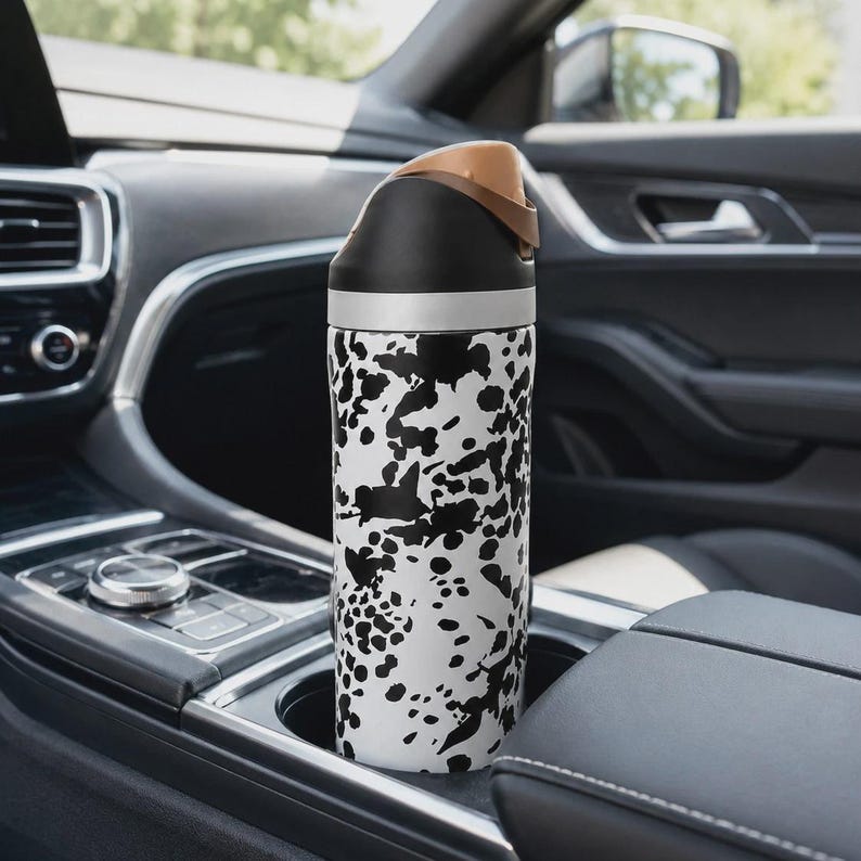 May include: A cylindrical insulated water bottle featuring a white base with an irregular black splatter pattern. The bottle has a black lid with a tan-colored flip-top spout and a silver-toned band separating the lid from the main body.