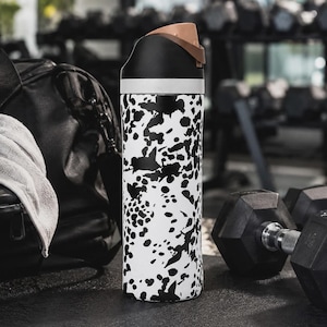 May include: A cylindrical water bottle featuring a white body with an irregular black splatter pattern. The bottle has a black lid with a light brown flip-top spout and a white band separating the lid from the main body of the container.