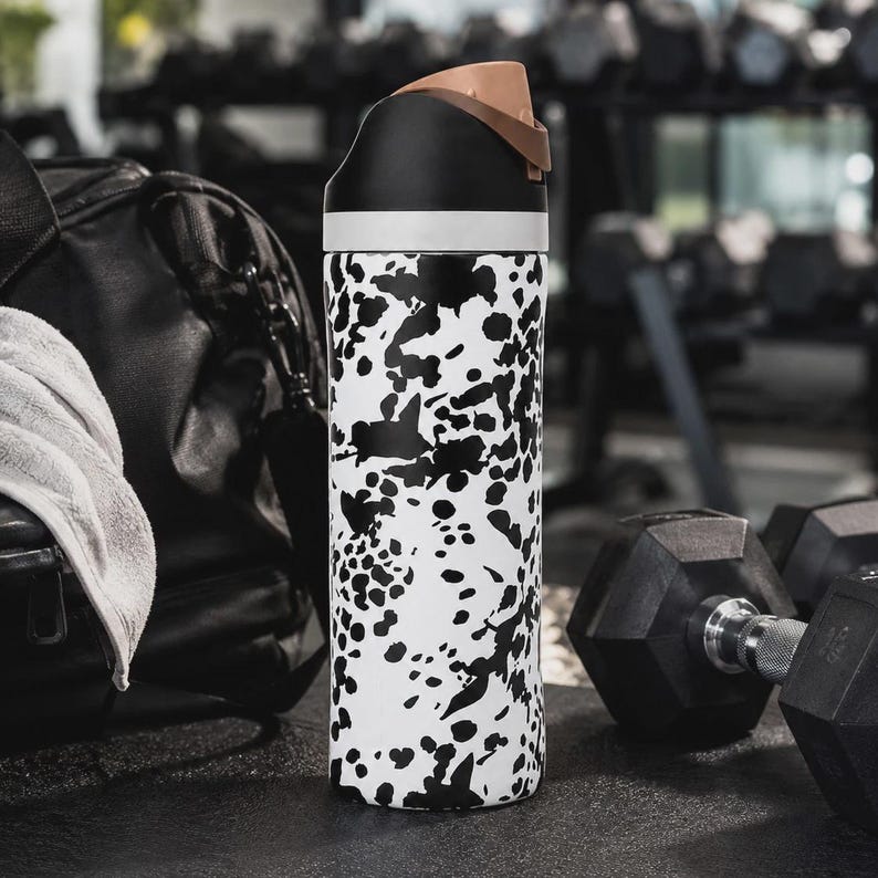 May include: A cylindrical water bottle featuring a white body with an irregular black splatter pattern. The bottle has a black lid with a light brown flip-top spout and a white band separating the lid from the main body of the container.