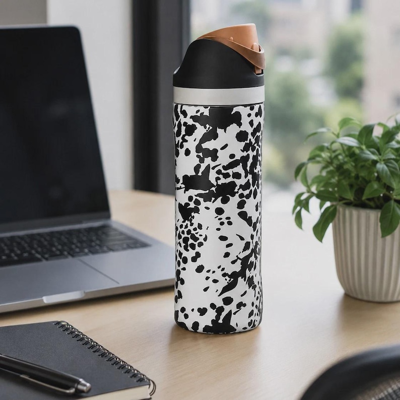May include: A cylindrical water bottle featuring a white base with an irregular black ink-blot or splatter pattern. The bottle has a black lid with a tan-colored flip-top spout and a silver-toned band separating the lid from the main body.