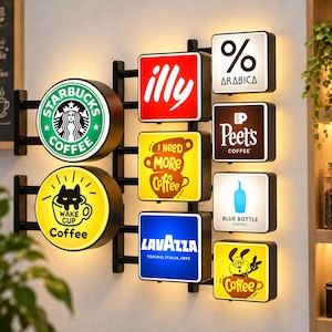 Könnte beinhalten: Eine Wandhalterung mit mehreren beleuchteten quadratischen und runden Schildern. Die Logos umfassen Starbucks, illy, % Arabica, Peet's Coffee, Blue Bottle Coffee und Lavazza sowie gelbe Schilder mit den Texten I Need More Coffee und Wake Cup.