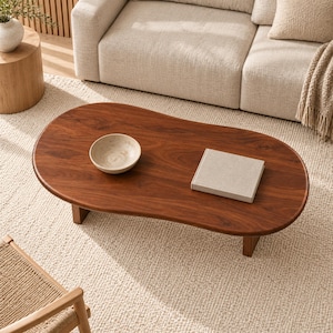 May include: A low, kidney-shaped coffee table made of dark brown wood with a smooth, polished surface. On the table sits a small, round, light-colored ceramic bowl and a square, light-colored book with a textured cover.
