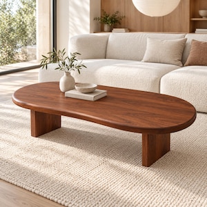 May include: A low-profile oval coffee table crafted from dark brown wood with a smooth, rounded edge surface. The table is supported by two thick, rectangular wooden legs and features a visible natural wood grain pattern throughout the top.