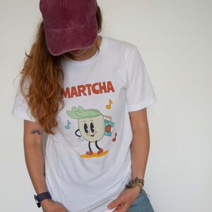 Puede incluir: Camiseta blanca con la palabra "MARTCHA" en naranja y un personaje de dibujos animados de cebolla con un radiocasete. La cebolla lleva zapatos rojos y está sobre una tabla de surf amarilla. También hay notas musicales en la camiseta.