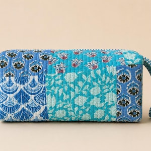 May include: A rectangular quilted pouch featuring a patchwork design of various blue and turquoise floral and fan-shaped patterns. The fabric has a stitched texture throughout, and a blue and white striped fabric handle is attached to the side.