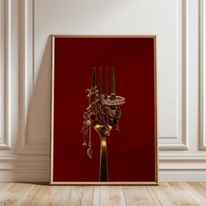 May include: A gold-toned metal fork centered against a deep red background, with a collection of gold-colored jewelry including chains, a ring with clear stones, and a small leaf-shaped charm draped over and tangled around the tines of the fork.