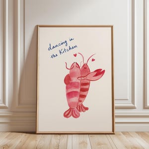 May include: A framed wall art print featuring a watercolor illustration of two red lobsters dancing together. The lobsters have small red hearts above them. The text "dancing in the Kitchen" is written in blue cursive script above the lobsters.