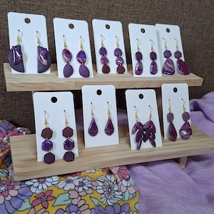 Op de afbeelding: Een collectie van tien paar oorbellen in paarse tinten, gepresenteerd op witte kaarten op een houten standaard met twee niveaus. De oorbellen hebben verschillende vormen, waaronder cirkels, zeshoeken en druppels, met gemarmerde en effen afwerkingen.