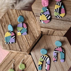 Peut inclure: Quatre paires de boucles d'oreilles pendantes en pâte polymère. Chaque paire présente une combinaison unique de formes géométriques aux couleurs vives, notamment rose, bleu, jaune, vert et violet, avec des motifs rayés noirs et blancs.