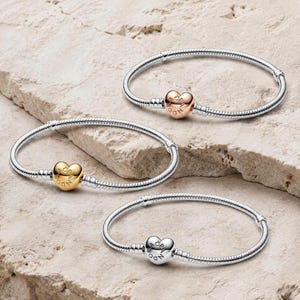 Könnte beinhalten: Drei Schlangenketten-Armbänder, jedes mit einem herzförmigen Verschluss. Ein Armband hat ein roségoldfarbenes Herz, eines ein goldfarbenes Herz und eines ein silberfarbenes Herz. Jeder Herzverschluss ist mit dem Wort Pandora graviert.