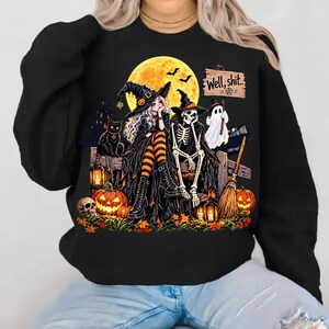 May include: A black long-sleeve sweatshirt featuring a graphic print of a witch and a skeleton sitting on a wooden fence under a full moon. The design includes a black cat, a ghost, pumpkins, lanterns, a broom, and the text Well, shit..