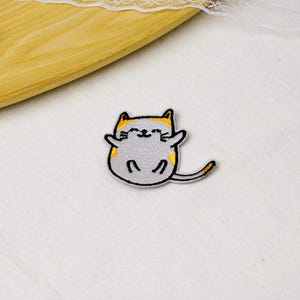 May include: An embroidered patch featuring a round, light gray cat with yellow ears and a yellow-tipped tail. The cat has a black stitched outline, closed eyes, and a small smile, with its arms outstretched to the sides in a reclining position.