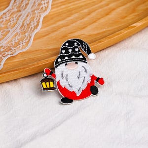 May include: An embroidered patch featuring a gnome with a long white beard, wearing a black hat with white heart patterns. The gnome has a red body, black boots, and red mittens, and is holding a yellow and black lantern in one hand.