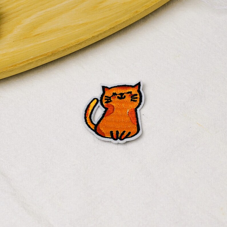 May include: An embroidered iron-on patch featuring a small, orange tabby cat with a simple black outline. The cat is depicted in a sitting position with its tail curved upward, showcasing detailed orange thread work and a friendly, minimalist facial expression.
