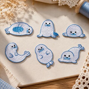 May include: A set of six embroidered patches featuring white harp seal pups in various poses. Each patch is outlined in blue thread, with black eyes, blue noses, and stitched whiskers. The seals are depicted lying down, sitting upright, and swimming.
