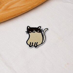 May include: An embroidered patch featuring a cartoon cat with a dark brown head and ears, a light beige body, and a curved tail. The cat has a simple stitched face with closed eyes and whiskers, outlined in white thread against a white background.