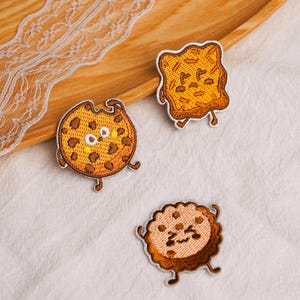 May include: Three embroidered fabric patches featuring anthropomorphic food characters. One patch is a round chocolate chip cookie with a bite taken out, one is a square piece of toast, and one is a round scalloped-edge cookie with a smiling face.