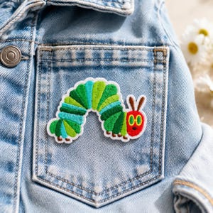May include: An embroidered patch featuring a colorful caterpillar with a red head, yellow eyes, and a segmented body in shades of green and teal. The patch is attached to the front pocket of a light blue denim jacket with a silver metal button.