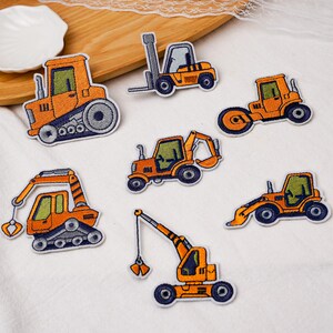 May include: A collection of seven embroidered patches featuring orange construction vehicles, including a bulldozer, forklift, steamroller, backhoe, excavator, and crane, all with dark blue wheels and green-tinted windows on a white fabric surface.