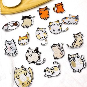 May include: A collection of fifteen embroidered cat patches in various poses and colors, including orange, grey, white, and brown tabby patterns. Each patch features a cartoon-style feline with stitched outlines and expressive facial features.