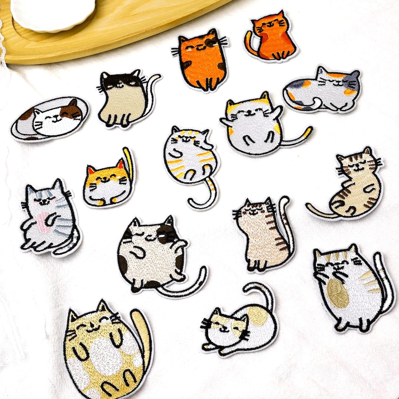 May include: A collection of fifteen embroidered cat patches in various poses and colors, including orange, grey, white, and brown tabby patterns. Each patch features a cartoon-style feline with stitched outlines and expressive facial features.