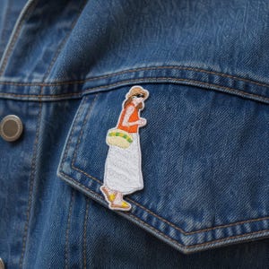 May include: An embroidered patch featuring a figure wearing a tan sun hat, sunglasses, an orange sleeveless top, and a long white skirt. The figure carries a yellow and green striped bag and wears yellow sandals, all attached to a blue denim jacket.