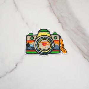May include: An embroidered patch shaped like a camera with a tan body, green top, and a large circular lens. The lens features a landscape scene with a red sun, orange sky, blue mountains, and a black silhouette of a tree. An orange strap hangs down.