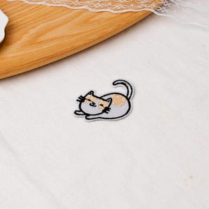 May include: An embroidered iron-on patch featuring a small, light gray cat lying down with its tail curled upward. The cat has a tan patch on its head and back, with black stitched details for its eyes, nose, mouth, and whiskers, outlined in white.