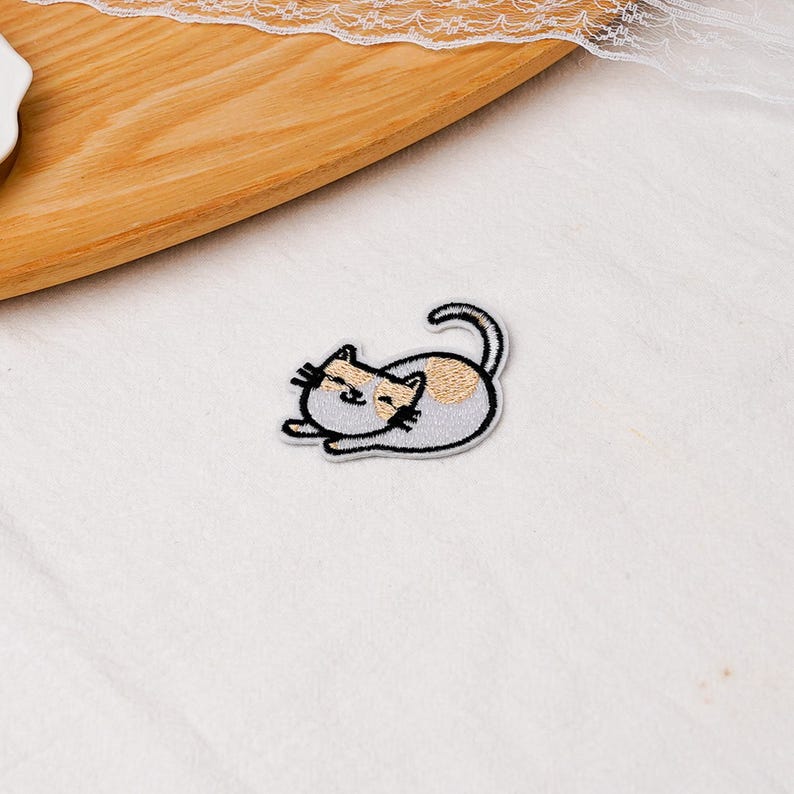 May include: An embroidered iron-on patch featuring a small, light gray cat lying down with its tail curled upward. The cat has a tan patch on its head and back, with black stitched details for its eyes, nose, mouth, and whiskers, outlined in white.