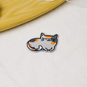 May include: An embroidered iron-on patch featuring a calico cat in a resting position. The cat has light gray fur with patches of orange and dark gray, closed eyes, and a small smile. The design is outlined in black thread with a white border.