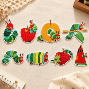 May include: A collection of eight embroidered patches featuring a green caterpillar with a red head in various poses, including crawling, eating a red apple, an orange, a strawberry, a slice of watermelon, and resting on a brown tree branch.